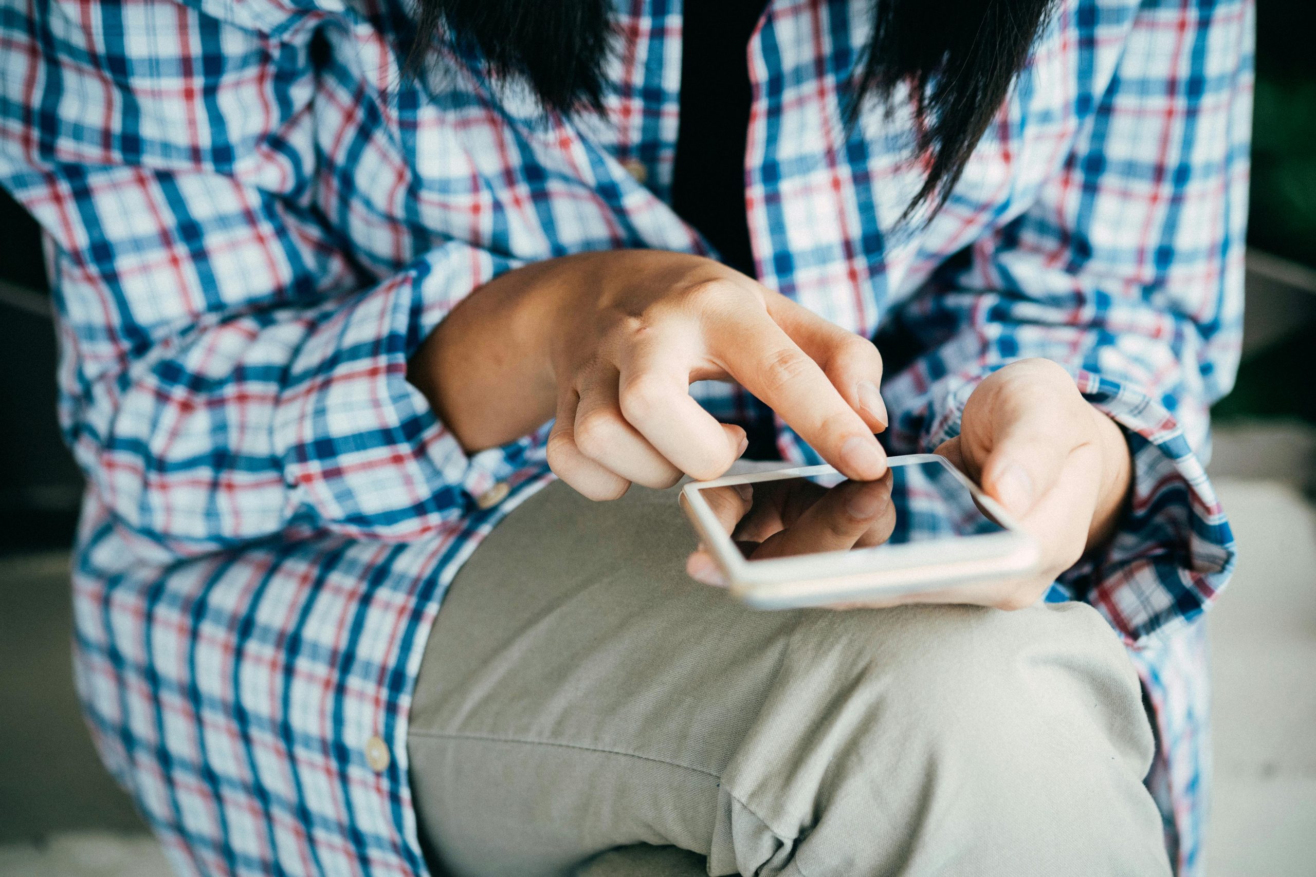 Woman's hands using a smartphone, wearing plaid shirt, indoors. Close-up shot highlighting touch interaction.