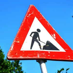 A red and white roadwork warning sign with a silhouette figure, set against a clear blue sky.